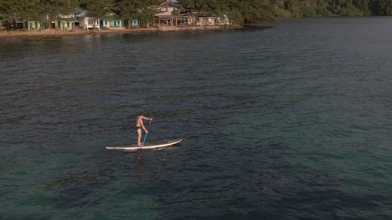 vista aérea de un hombre haciendo ejercicio en una tabla de remo en aguas turquesas tropicales claras, con costa rocosa y bungalows estilo mochilero en koh chang tailandia