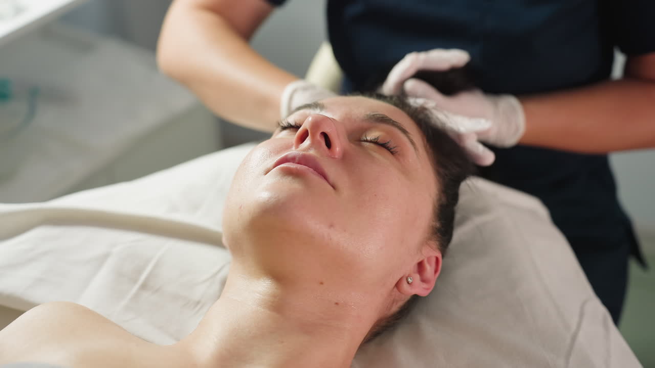 Beauty technician in gloves gently packs client black hair then wraps head with white cotton towel while recipient peacefully closes eyes during spa treatment in bright modern treatment room