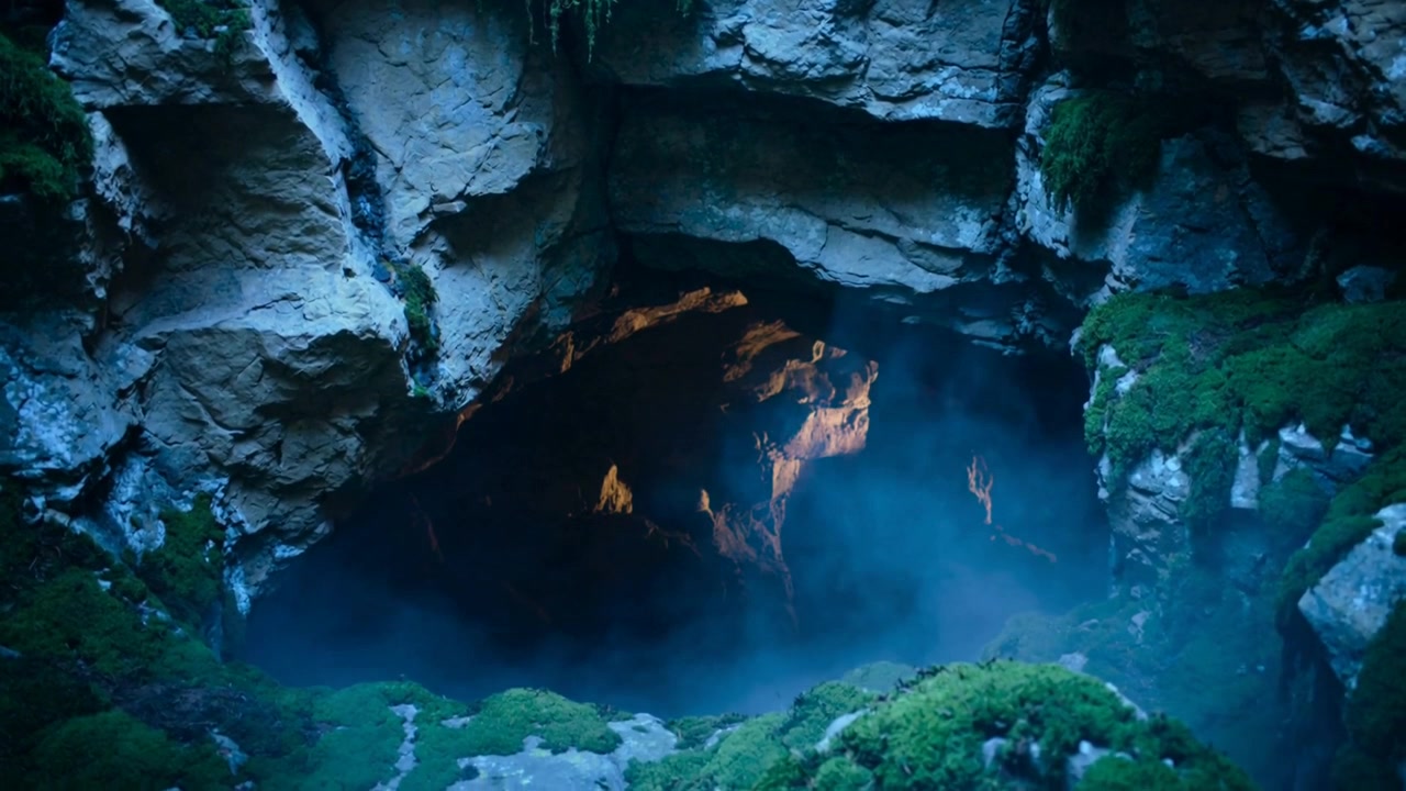 Swirling bluish mist beginning revealing cavern mouth at mossy cliff, exposing warm orange glow