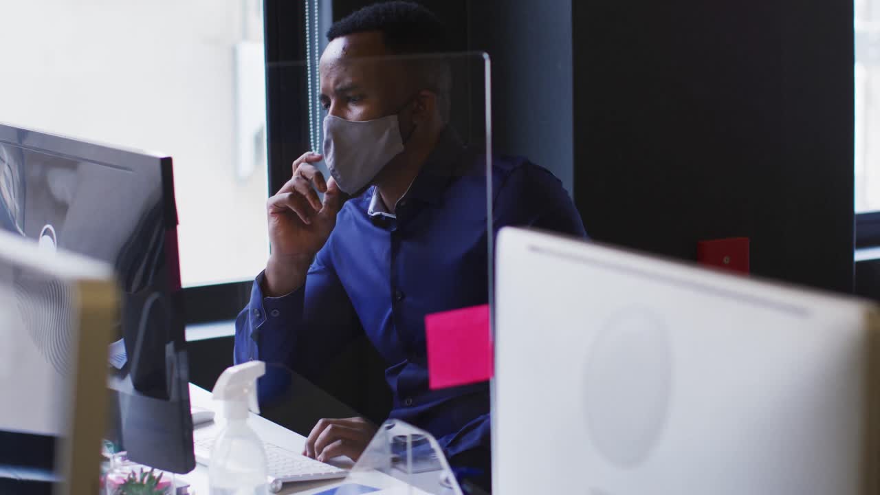 African american man wearing face mask using computer while sitting on his desk at modern office