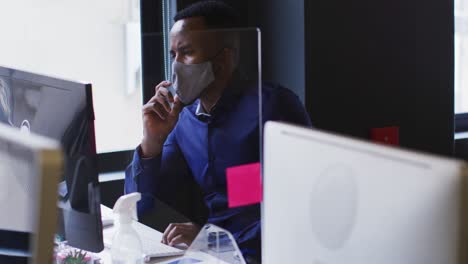 african american man wearing face mask using computer while sitting on his desk at modern office