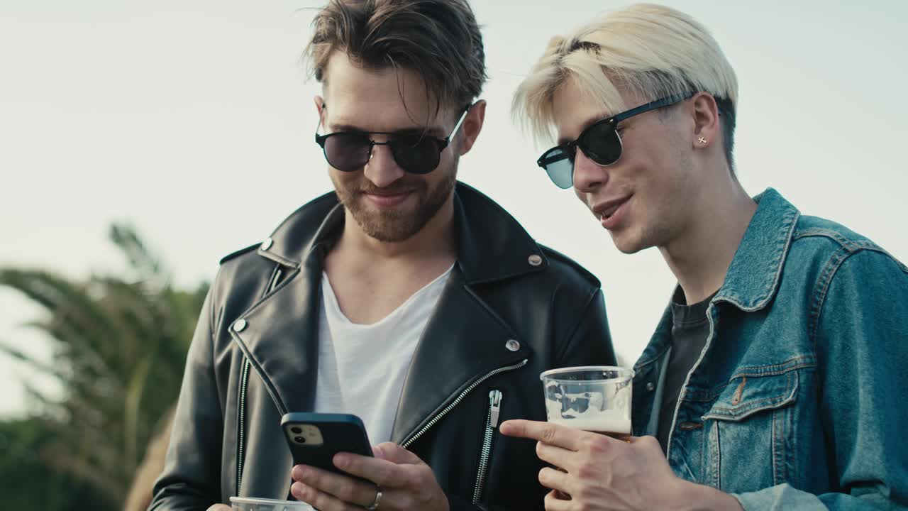 Due giovani uomini caucasici che navigano al telefono e si divertono a un festival musicale mentre bevono birra