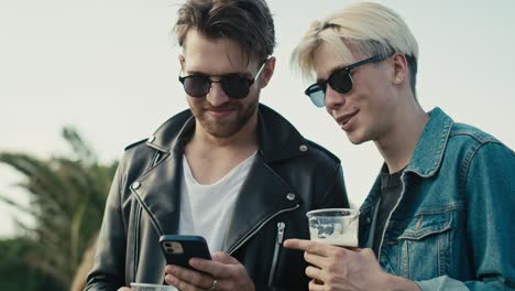 two young caucasian men browsing phone and having fun on music festival while drinking beer.