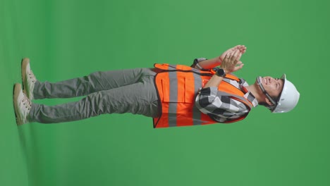 full body side view of asian male engineer with safety helmet smiling and clapping his hands while standing in the green screen background studio