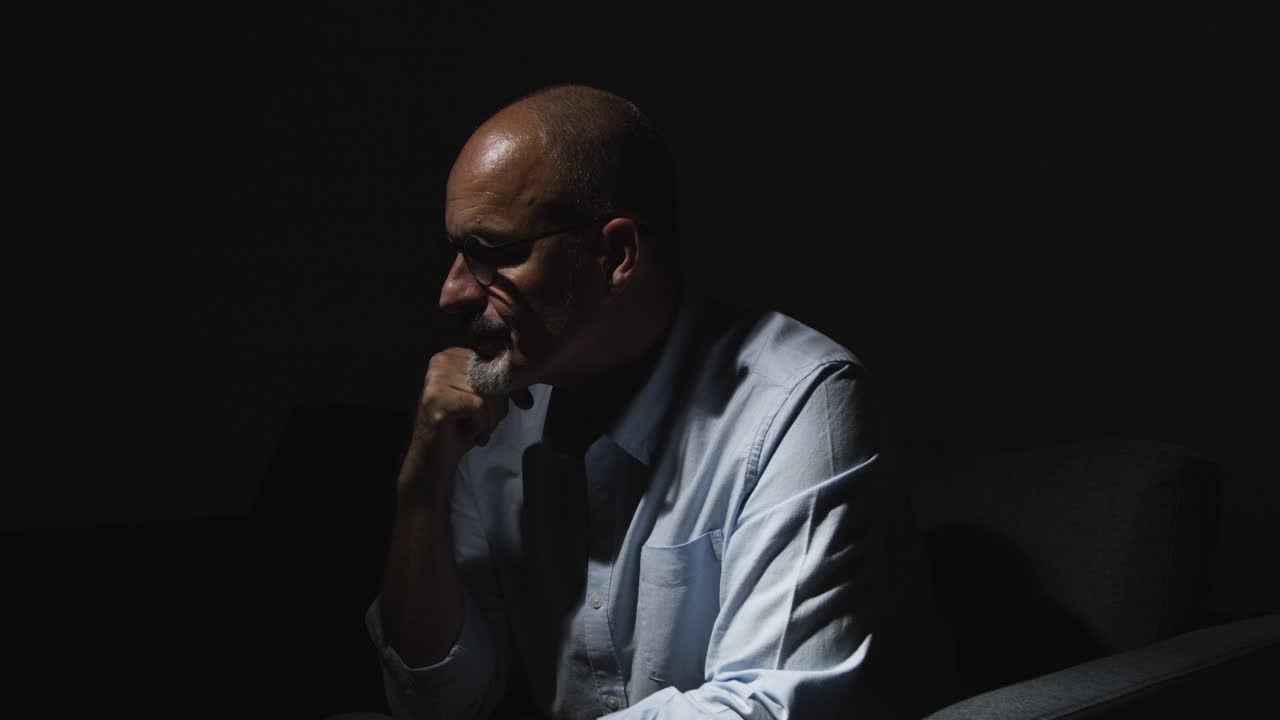Depressed Mature Man Struggling With Mental Health Sitting In Darkness On Sofa With Low Key Lighting