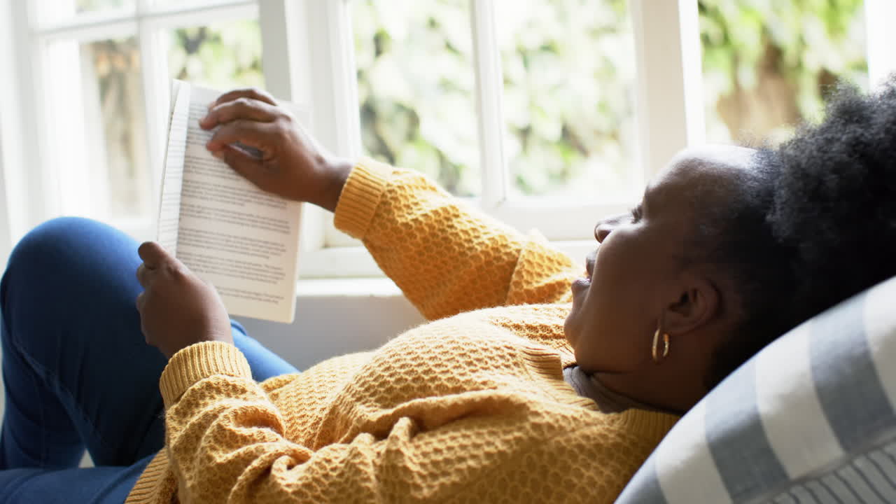 Happy african american senior woman lying, reading book and smiling in sunny room, slow motion
