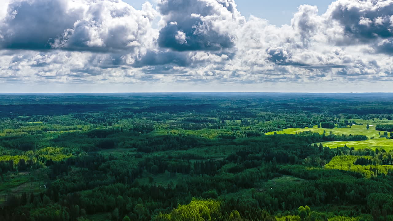 A static aerial timelapse of fast-moving clouds and their shadows sweeping across a vast and beautiful landscape of dense green forests and fields on a summer day