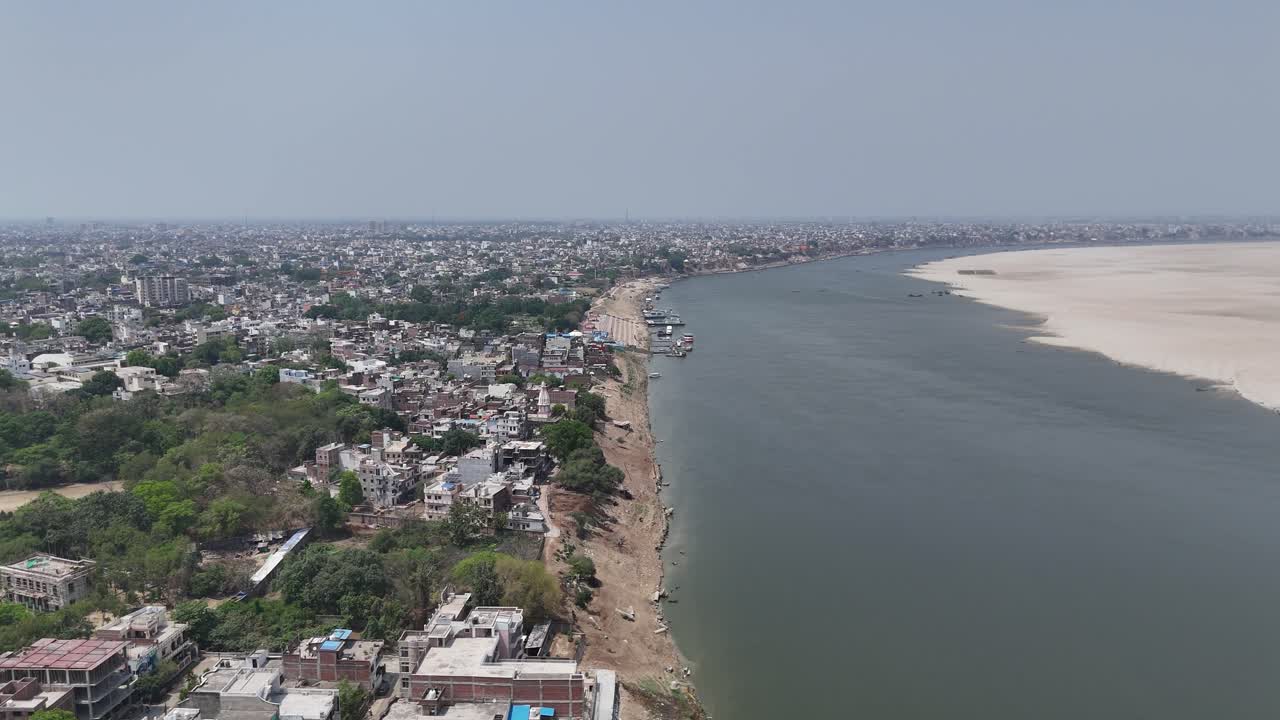 A panoramic aerial view of Varanasi's riverfront, where the timeless Ganges flows beside a dense tapestry of historic buildings and bustling markets, reflecting the city's ancient spirit.