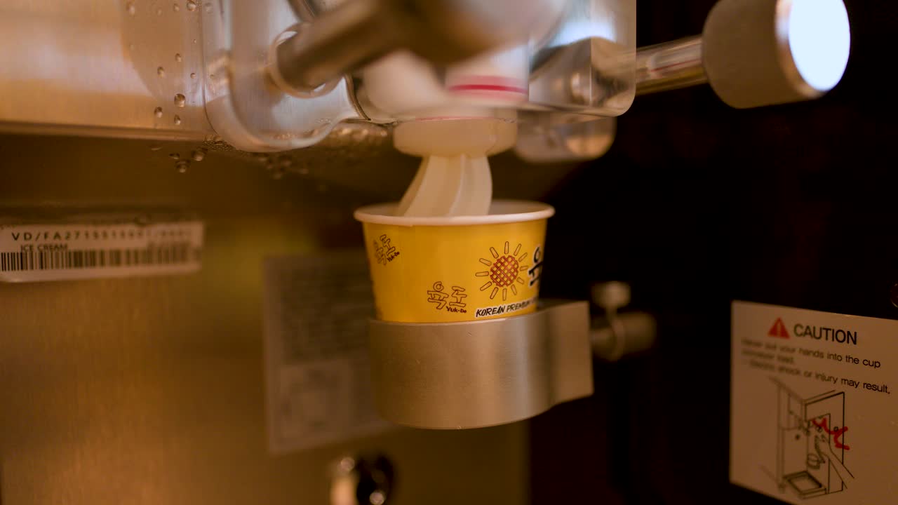 Soft serve ice cream dispensed into a paper cup by machine, warm lighting, close-up view