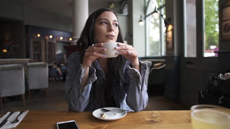 Attraktive-Latina-Brünette,-Die-Am-Tisch-Sitzt-Und-Einen-Kaffee-Trinkt,-Wartet,-Nachdenkt-Und-Nach-Draußen-Schaut