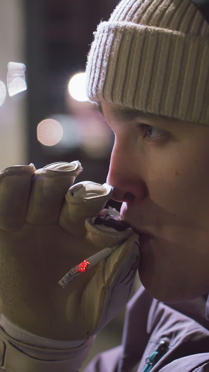 헤드폰과 장갑을 착용 한 후디를 입은 운동 선수의 근접 측면 뷰, 반사 표정으로 겨울 재을 입고 밤에 야외에서 담배를 피우고