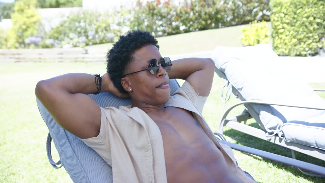 African-American man reclining on garden chaise in shirt feeling sun adjusting shades and settling