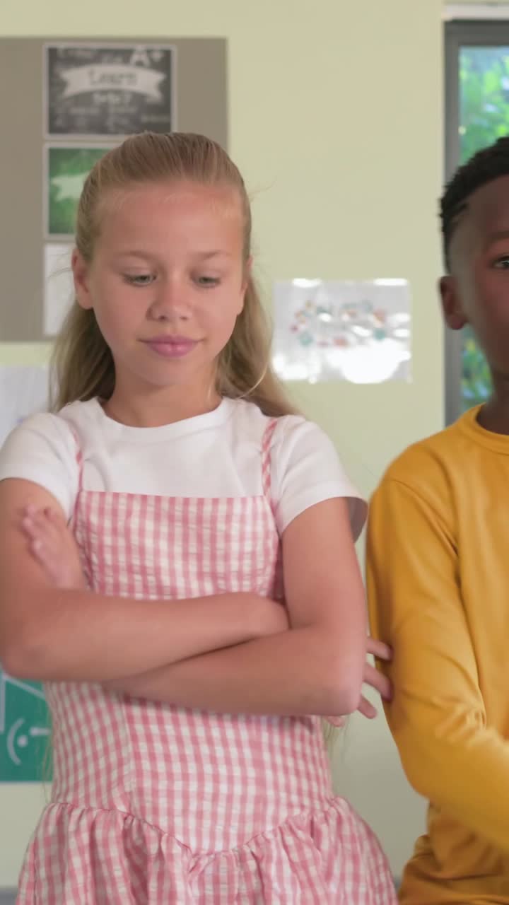 Vertical video: Entering left boy in gold shirt crossing arms leaning posing by board in class