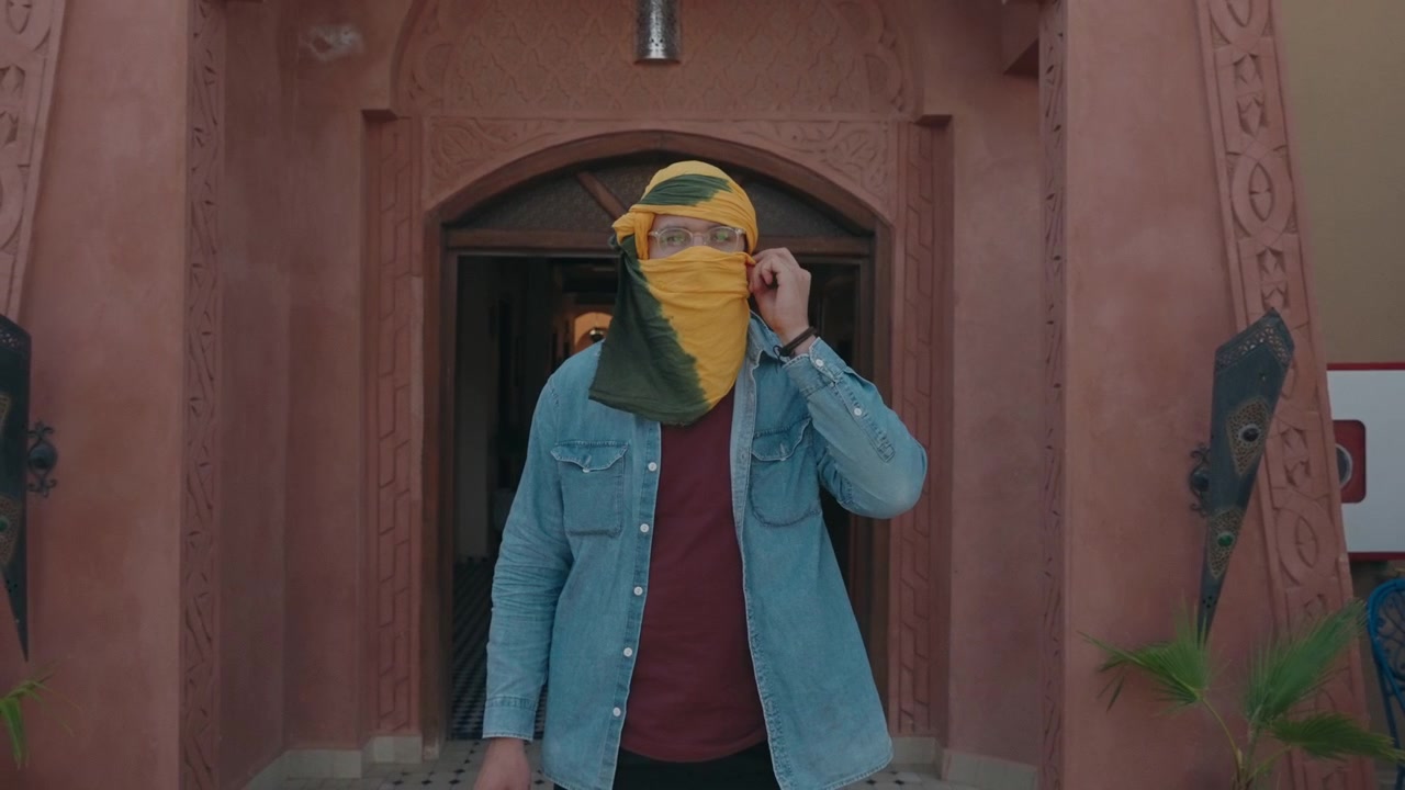 Giovane uomo che indossa una sciarpa giallo-verde sul viso e occhiali, in piedi sotto un arco di riad in terracotta intagliata.