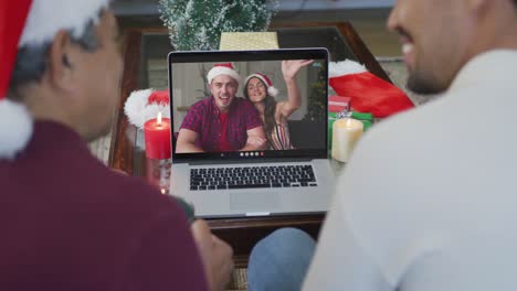 Smiling-biracial-father-and-son-using-laptop-for-christmas-video-call-with-couple-on-screen