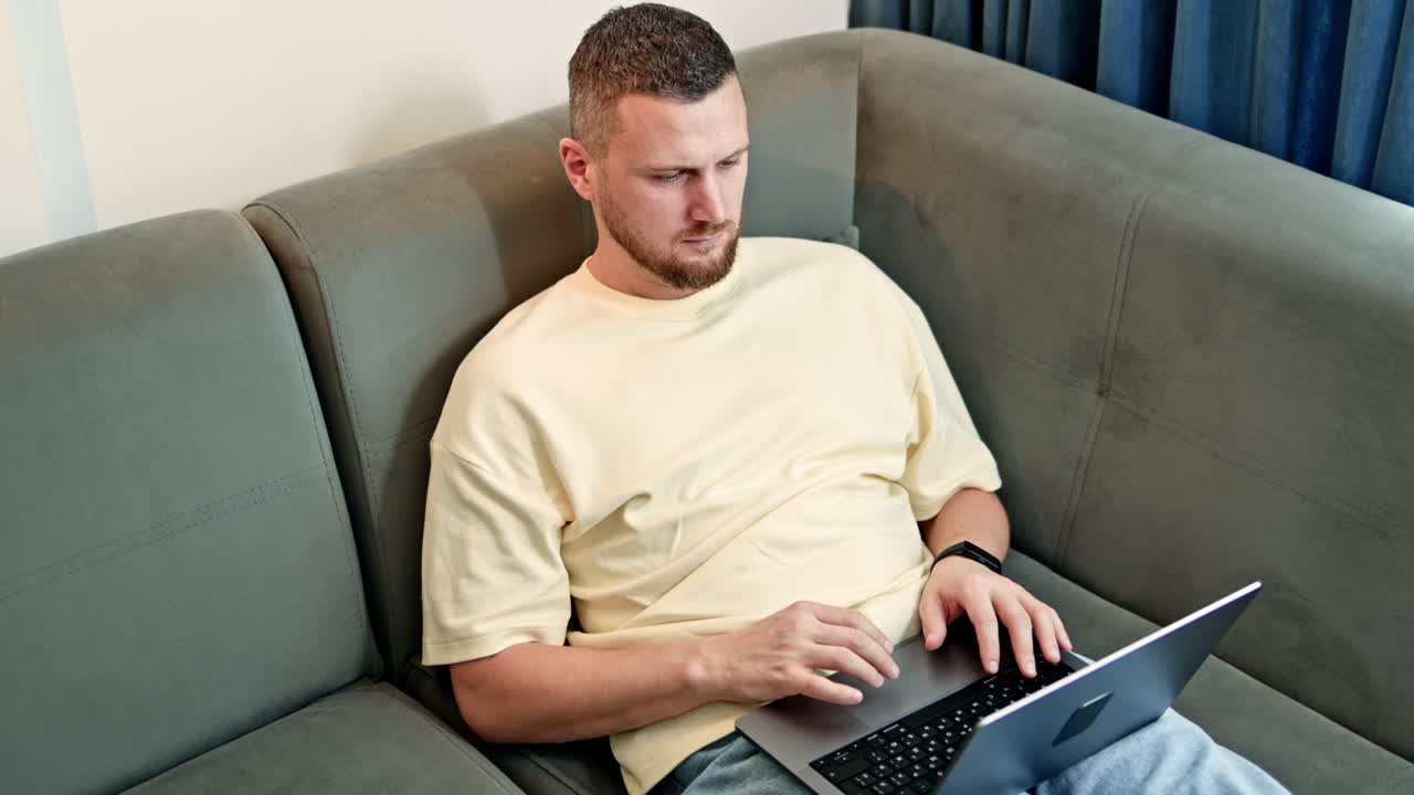 Mand sidder på grå sofa og bruger bærbar computer i et lyst rum om dagen