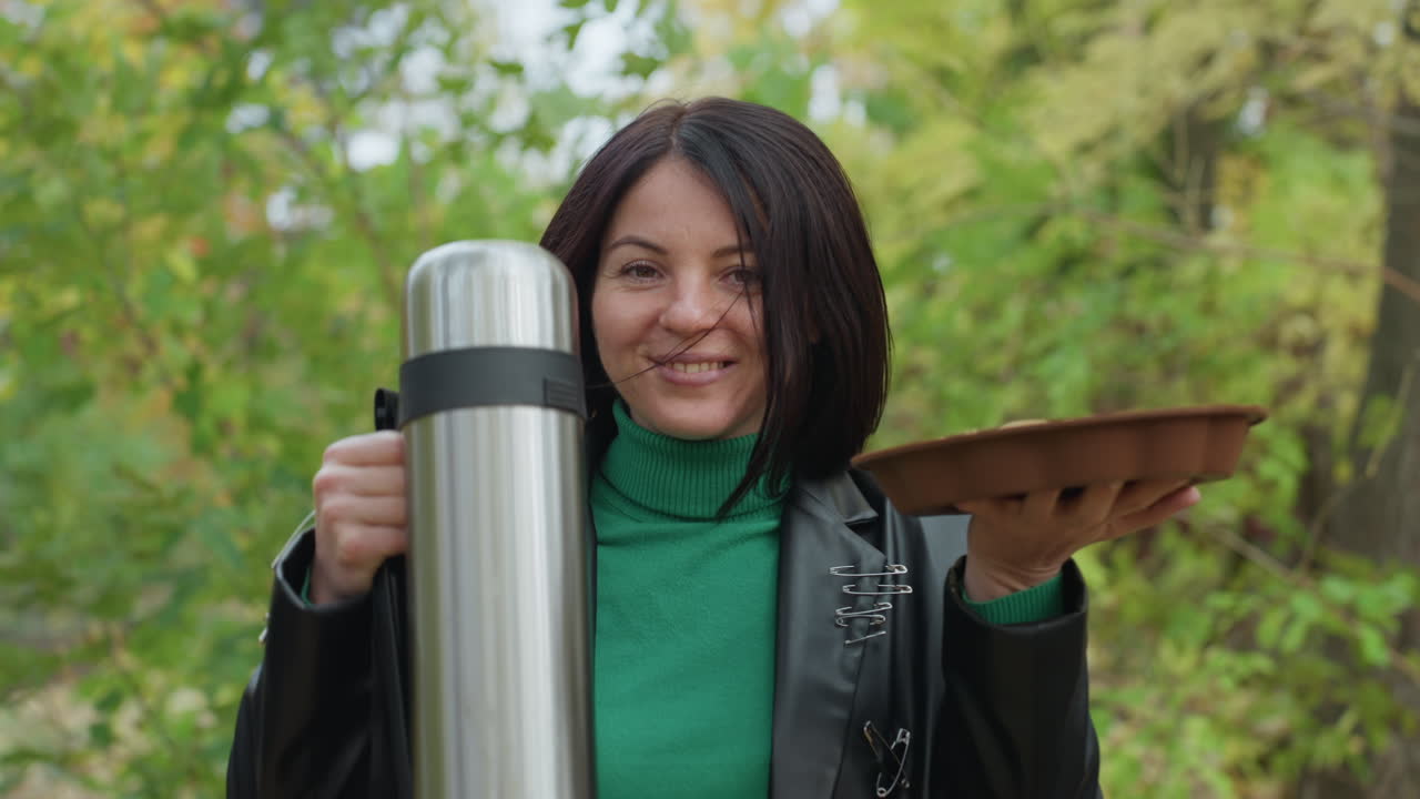 Donna di colore con giacca di pelle che tiene in mano un thermos, scena di picnic all'aperto in una foresta con vassoio di legno e borraccia in acciaio inossidabile, maglione verde, foglie autunnali, sorriso cordiale, atmosfera casual da rocker, gesto da padrona di casa sicura di sé