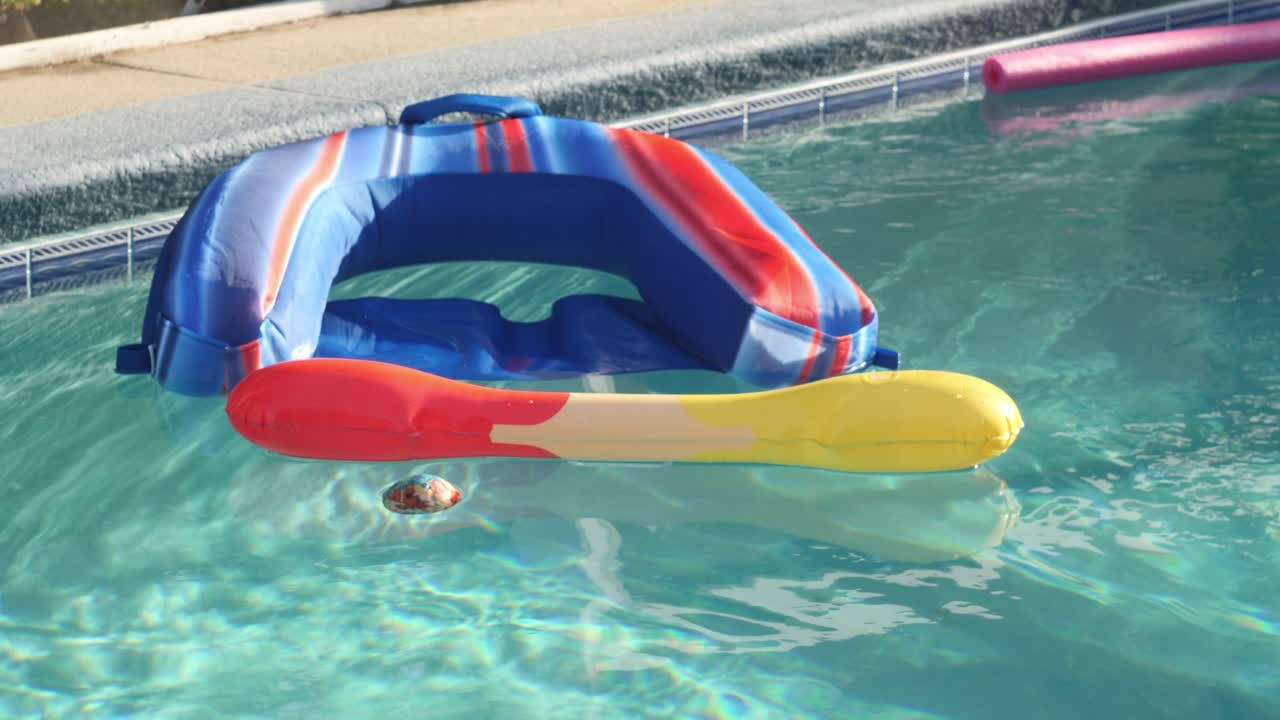 Pool noodle, blue floaty, ball, and stick with red and yellow ends (toys) floating in the pool in slow motion