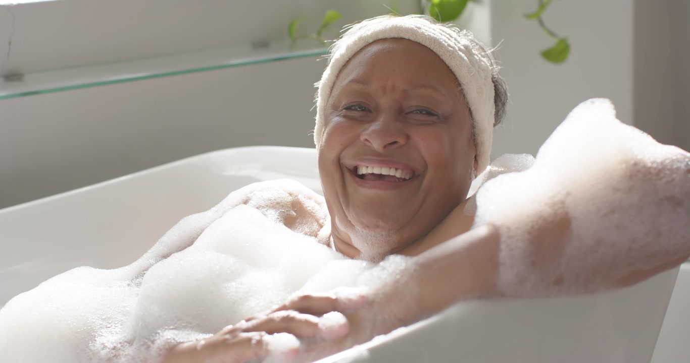 Senior African American woman in bubble bath with towel headband sunlight waking eyes open smiling