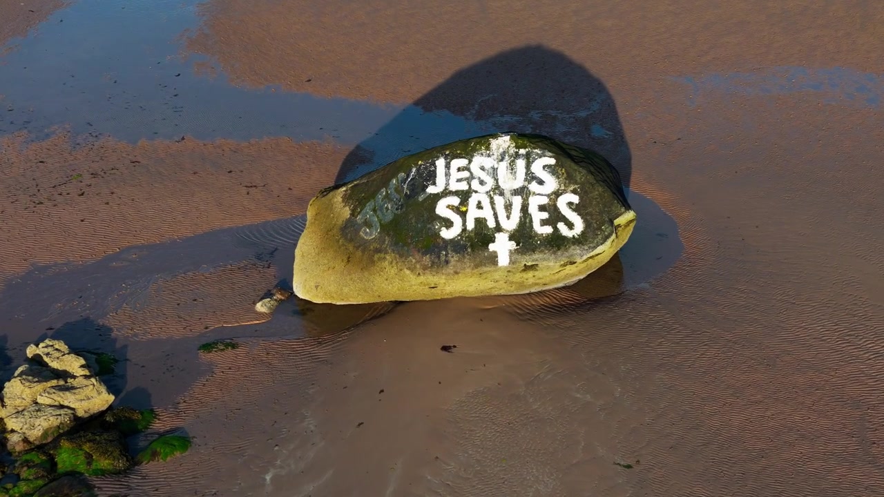Top-down drone dolly forward view, beach rock painted with the words “Jesus Saves” on the west coast of Scotland, showing a Christian message beside the shoreline at low tide