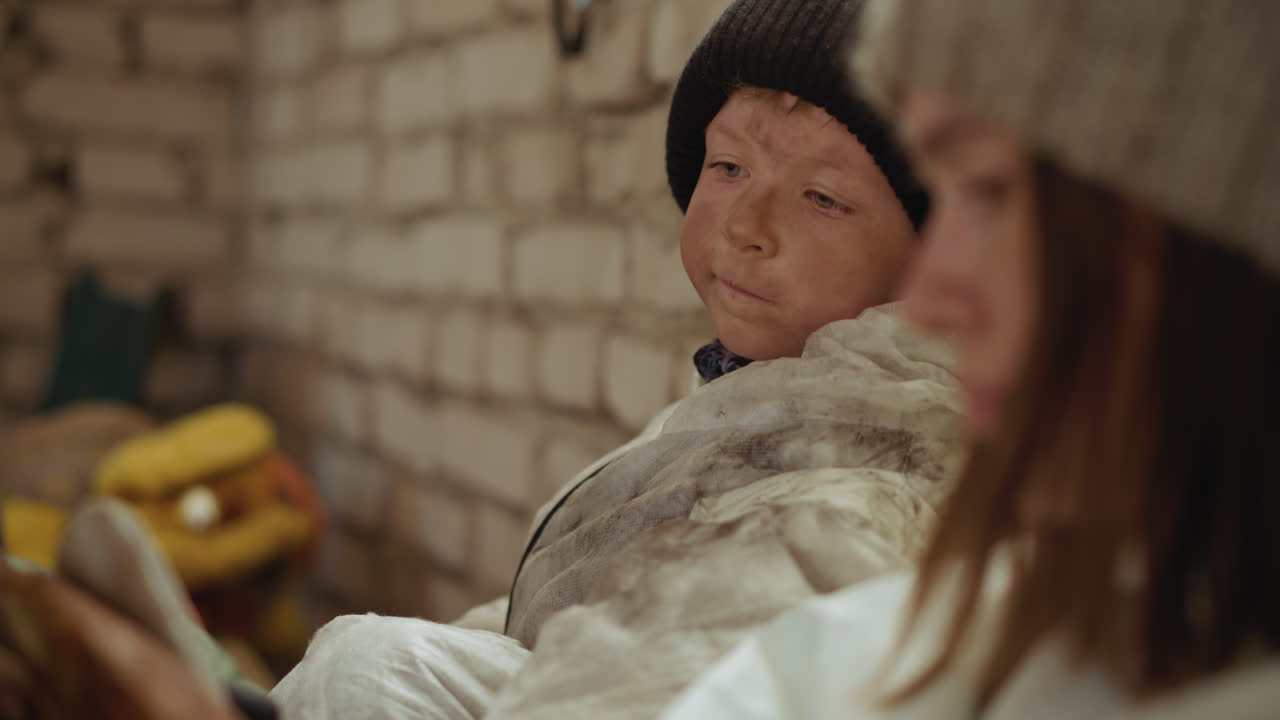 Worried young boy in dirty white protective suit and black knit cap peers sideways while resting against rough brick wall, partially leaning on leader beside