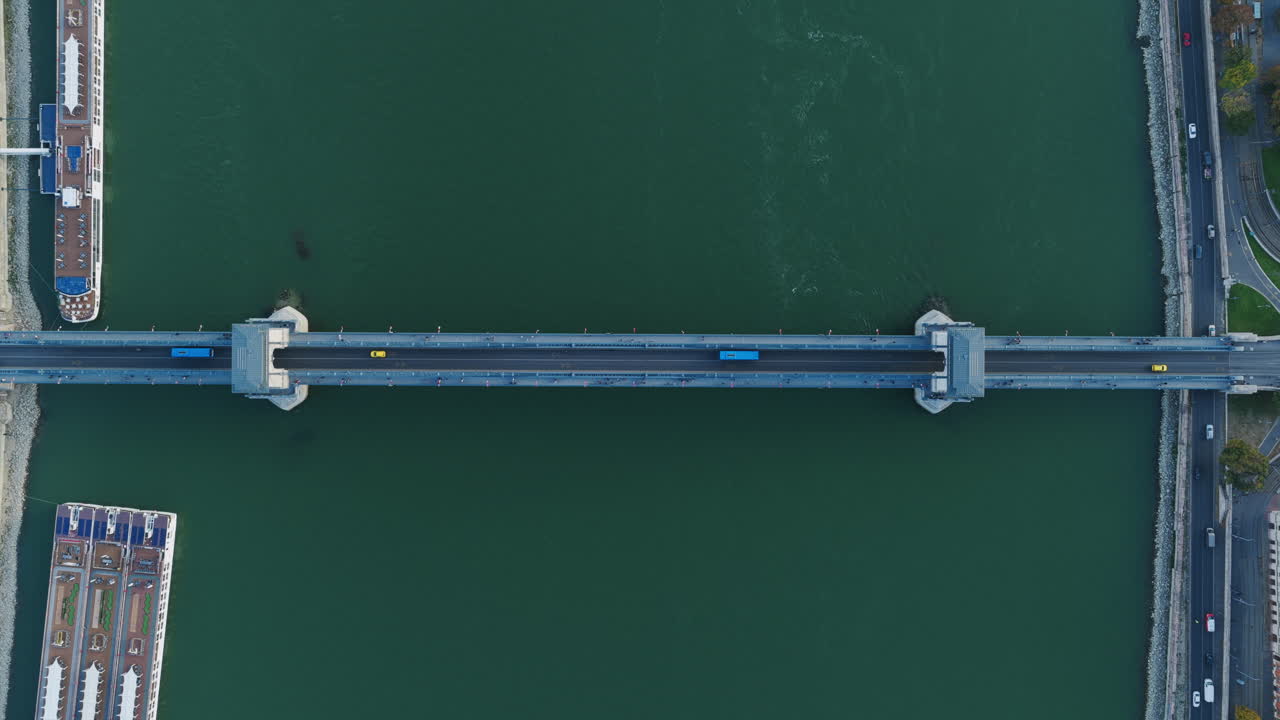 Aerial symmetry of a blue bridge with a yellow car and ferry below, highlighting Budapest’s urban geometry
