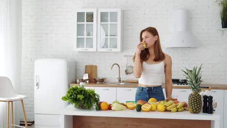 A beautiful sports girl at home in the kitchen eats a fresh apple. Table with different seasonal fruits. Vegetarian healthy food. Diet