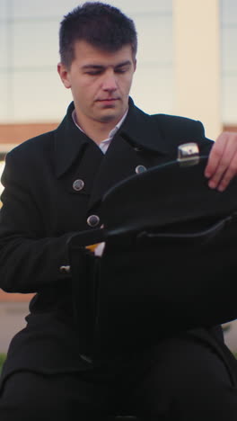 man in black suit sits on bench, opens briefcase in outdoor setting with modern building, parked car, and background featuring glass building reflecting light and surrounding structures