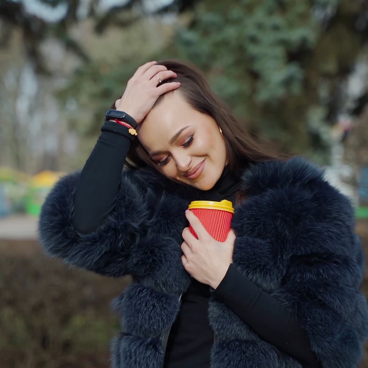 Portrait of a smiling woman. Attractive brunette girl in fur coat stands with plastic cup of coffee in the park and looking on camera. Slow motion.
