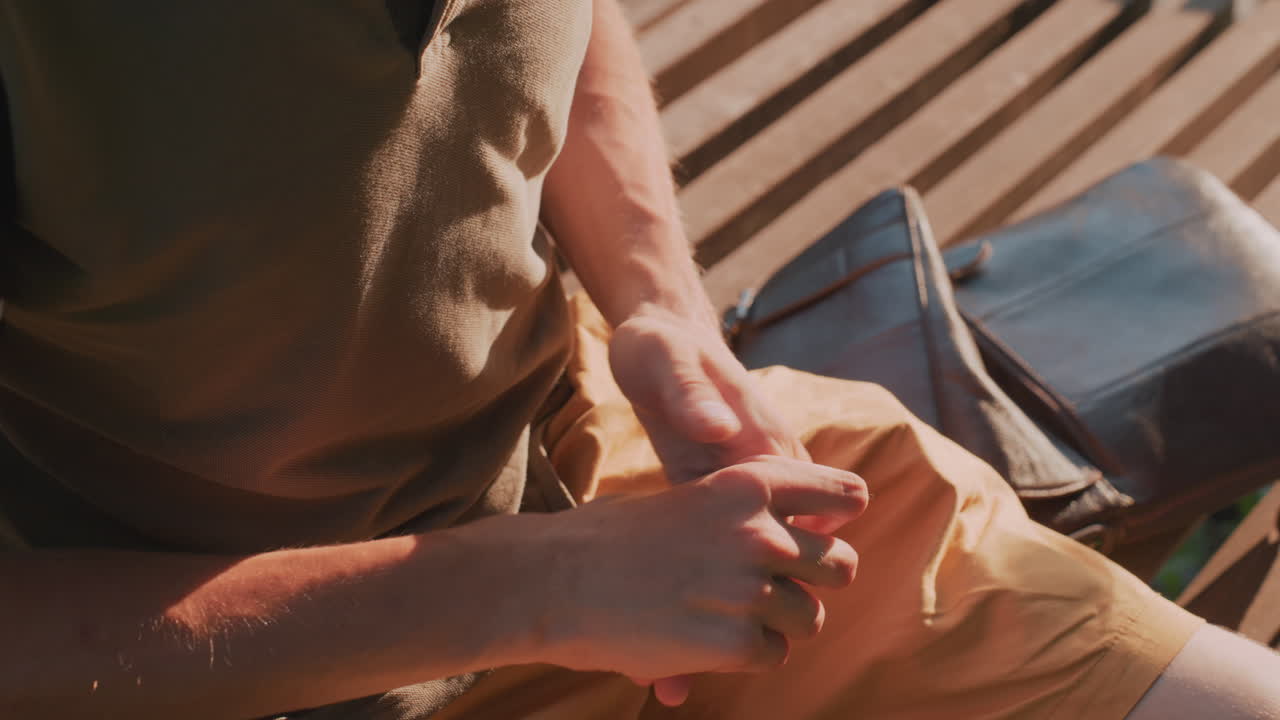 Man With Mobile, Man Seated On Bench Engaged With Device, Person Adjusting Belongings While Sitting In Park Setting, Male Individual Focused On Smartphone Amidst Warm Outdoor Park Environment
