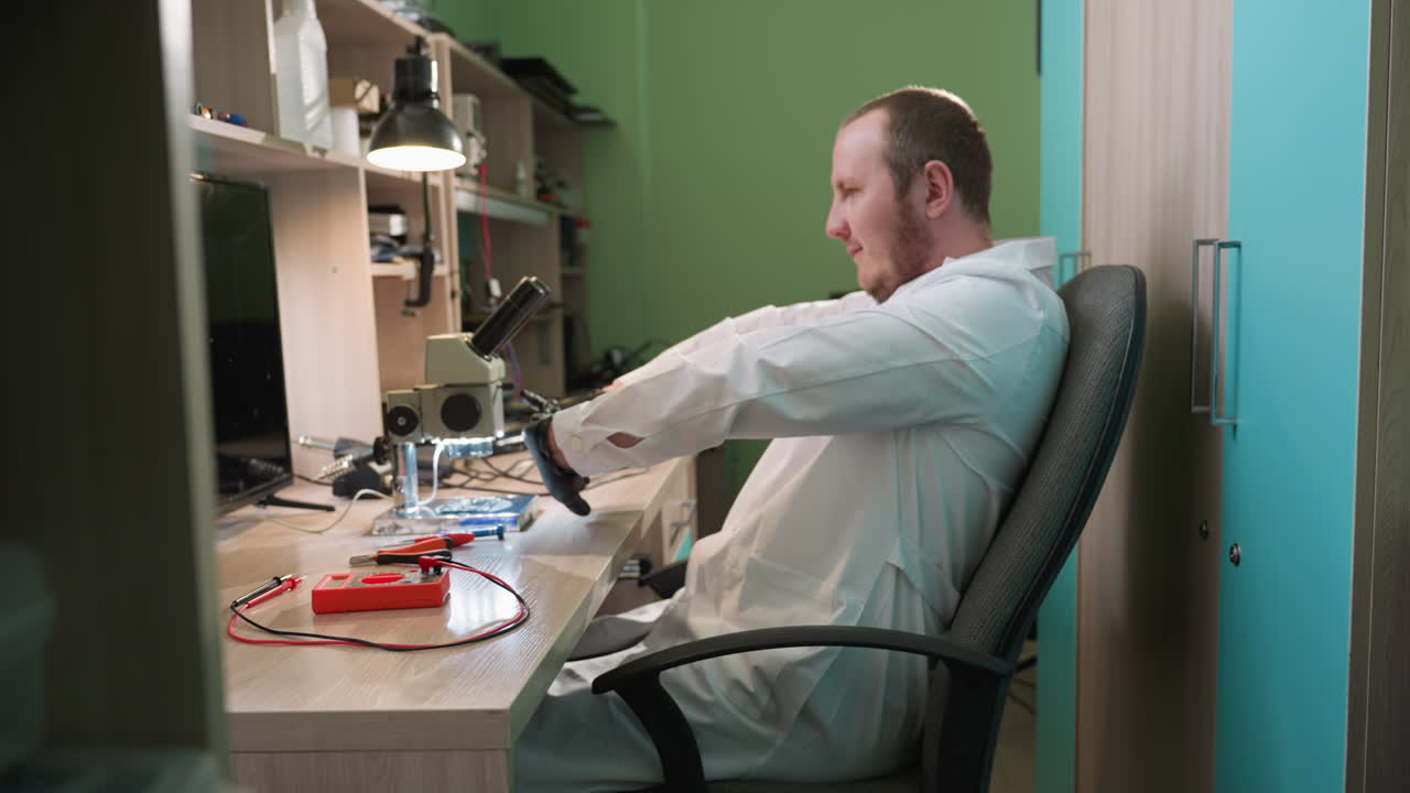 색 실험실 코트와 검은 장갑을 입은 기술자가 의자에 기대어 손을 펴고 현미경을 바라보면서 잠시 휴식을 취하는 근접한 모습