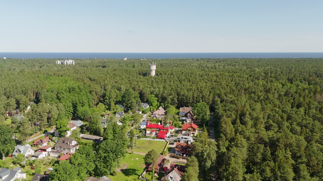 Aerial drone pull away reveals a lush pine forest in Jurmala, Latvia, with colorful residential houses, a distant water tower, and the Baltic Sea stretching across the background under clear skies