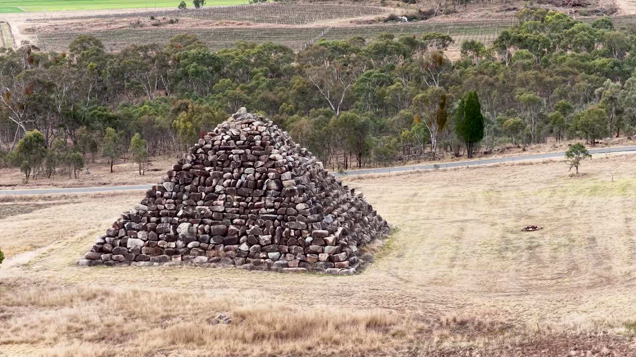 A stone pyramid stands in a dry, grassy field as the camera smoothly orbits, revealing surrounding trees and farmland under soft daylight