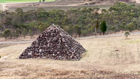 a stone pyramid stands in a dry, grassy field as the camera smoothly orbits, revealing surrounding trees and farmland under soft daylight