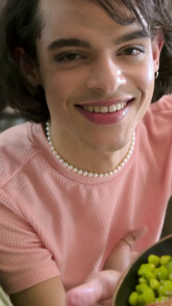 Vertical video: Speaking man in pink top presenting bowl in kitchen, offering edamame to friend