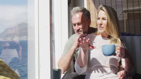 Happy-caucasian-mature-couple-drinking-coffee-and-talking-in-sunny-living-room