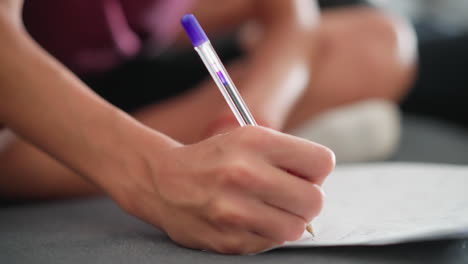 close up of young person writing with ballpoint pen while seated on sofa with slightly blurred background showing legs crossed and casual home environment