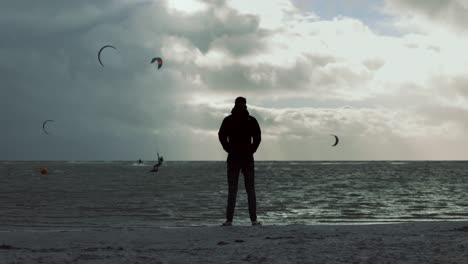 Mannsilhouette,-Die-Am-Ufer-Steht-Und-Kitesurfer-Im-Ozean-Mit-Dunklen-Wolken-Aufgrund-Von-Sturm-Francis-Beobachtet