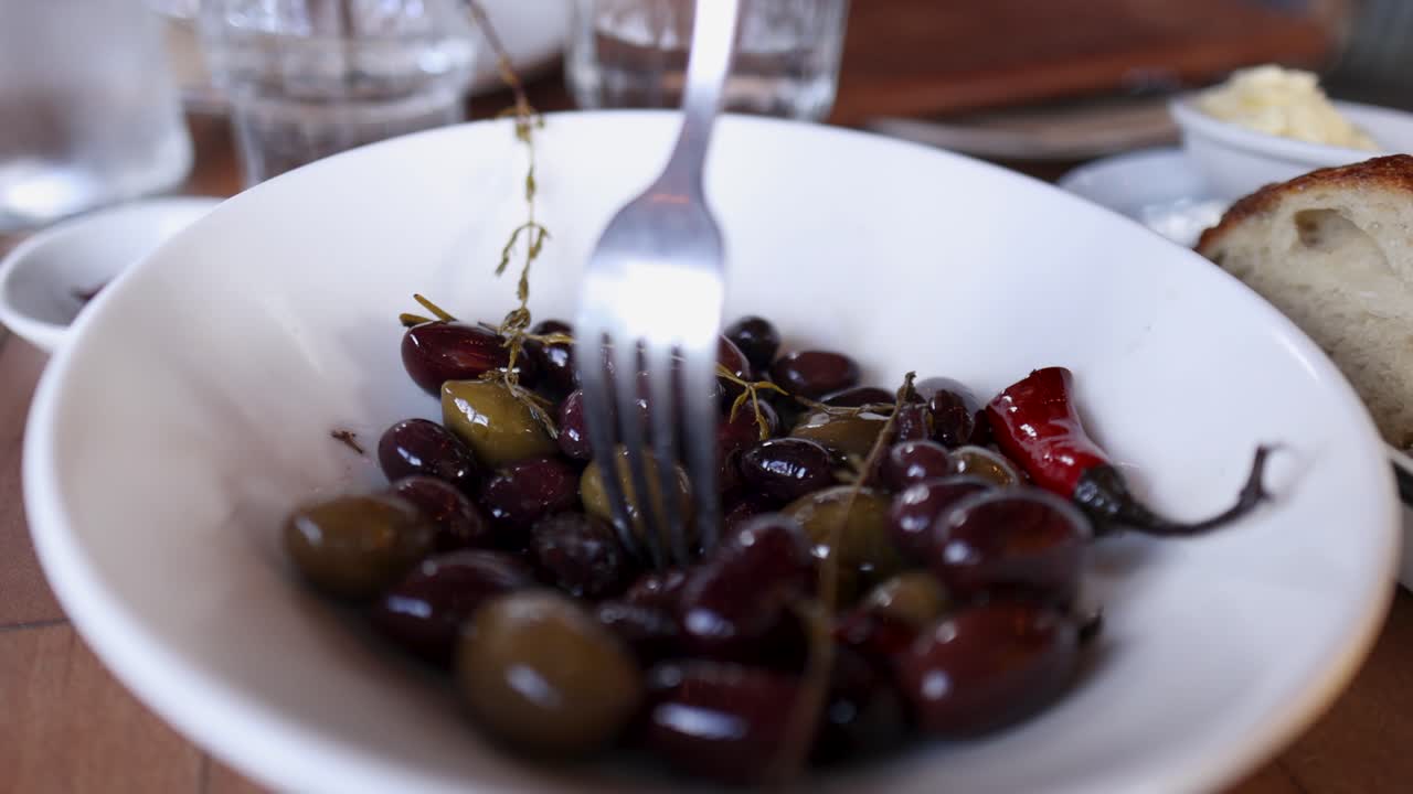 A hand picks marinated olives from a bowl on a wooden table in natural light, creating a casual dining atmosphere