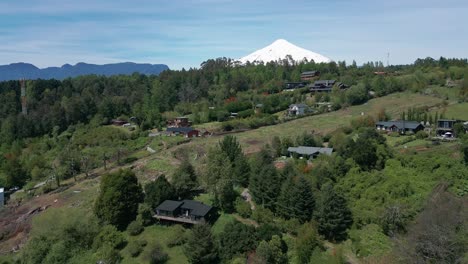 drone backward shot revealing hillside cabins and villas amid lush green forests with the snow-capped villarrica volcano dominating the background in southern chile