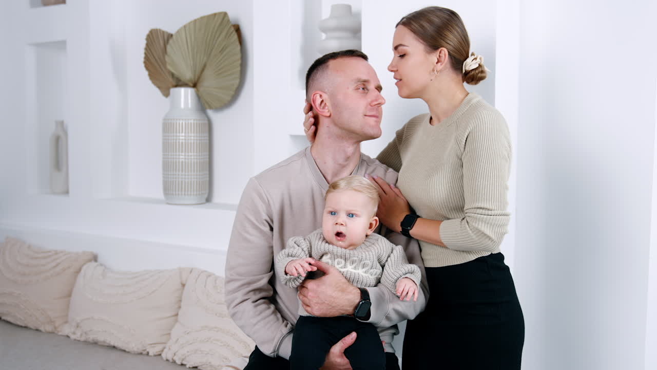 Loving Caucasian couple with an adorable baby. Mom and dad lean to each other, woman kisses her husband on the forehead tenderly.