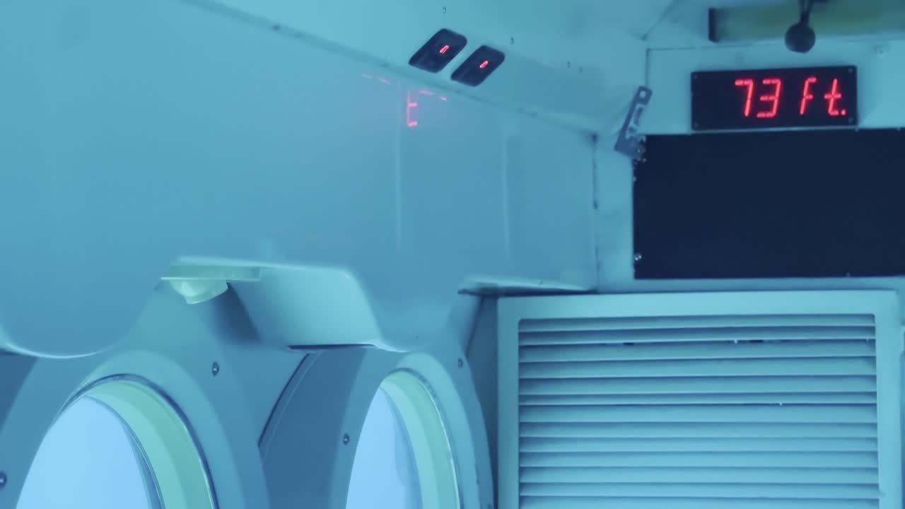 Cinematic panning shot from a row of portholes in a submarine to the depth gauge displaying 73 feet below the ocean's surface off the coast of Hawai'i
