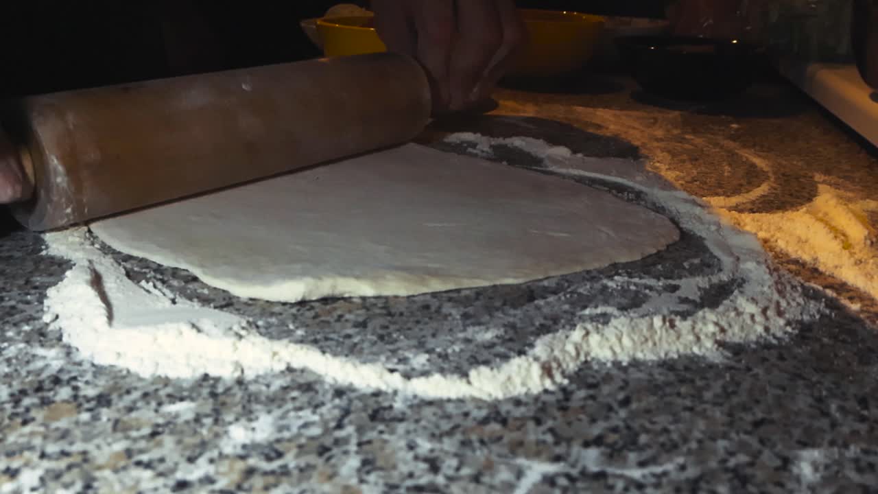 Chef or home cook using a rolling pin to make soft white and brown colored fluffy pizza douch on home kitchen countertop or table in slow motion with white colored fluffy flour around. Shallow depth