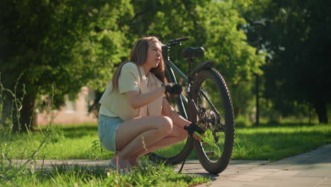 young woman in black gloves crouched by her bicycle, inflating its back tire, she appears tired but determine on a sunlit path. surrounded by vibrant greenery