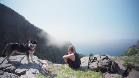 bearded male trekker resting sitting on cliff ledge eating a protein bar with his siberian husky dog beside him at mount donnamannen, norway