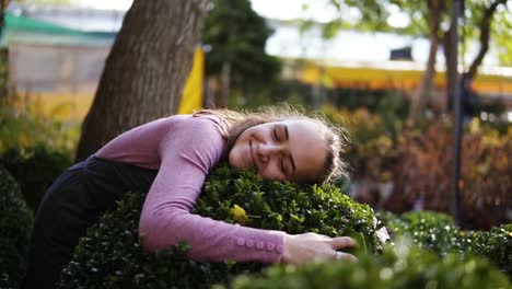 Florista-Sonriente-En-Delantal-Abrazando-Boj-Verde-En-El-Invernadero,-Sonriendo-Y-Mirando-A-La-Cámara.-Una-Mujer-Feliz-En-El-Invernadero-Ama-Los-Buxus-Y-Su-Trabajo.