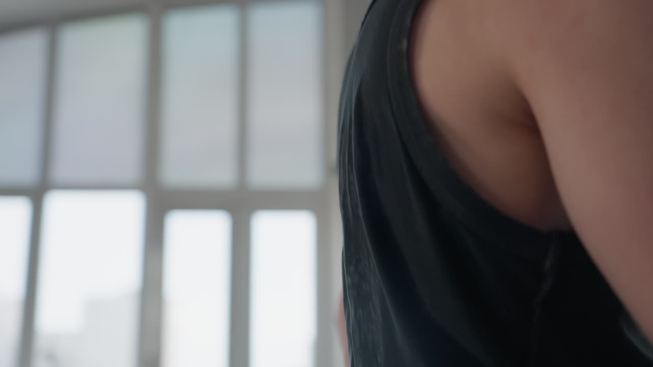 student in black vest walks to chair by large mirror and picks cream container from seat in minimalist studio with open security case on floor and tall window showing urban skyline