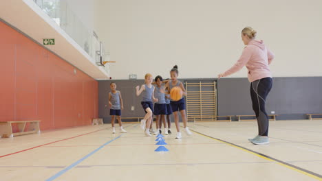 in school gym, children dribbling basketballs through cones with coach watching
