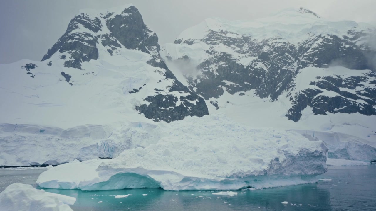 Picos oscuros irregulares se revelan sobre un vasto glaciar mientras los icebergs flotan a la deriva en las aguas turquesas del Océano Antártico.