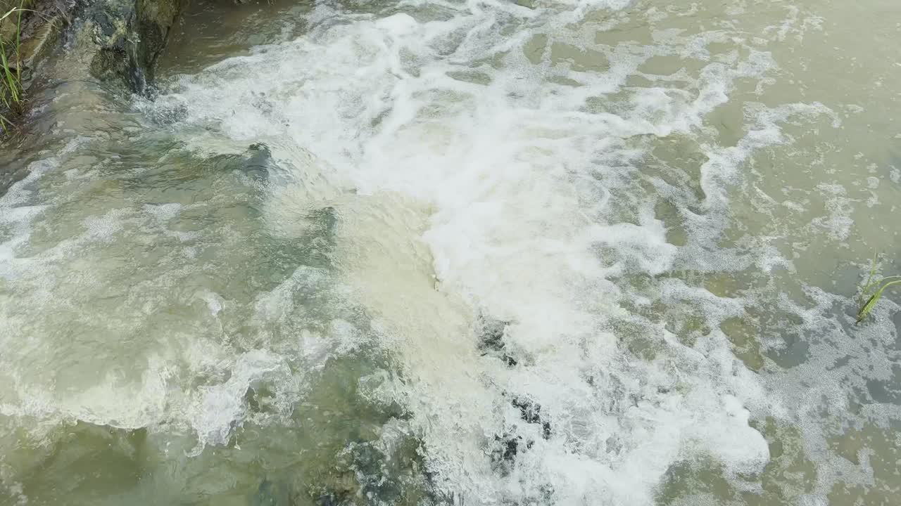A dynamic scene of turbulent, rushing water from a section of a river stream with a strong current that creates prominent areas of white, frothy foam and splashes